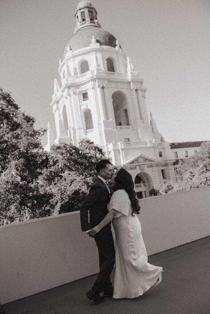 Black and white photo of a couple kissing with the Pasadena City Hall rotunda behind them.
