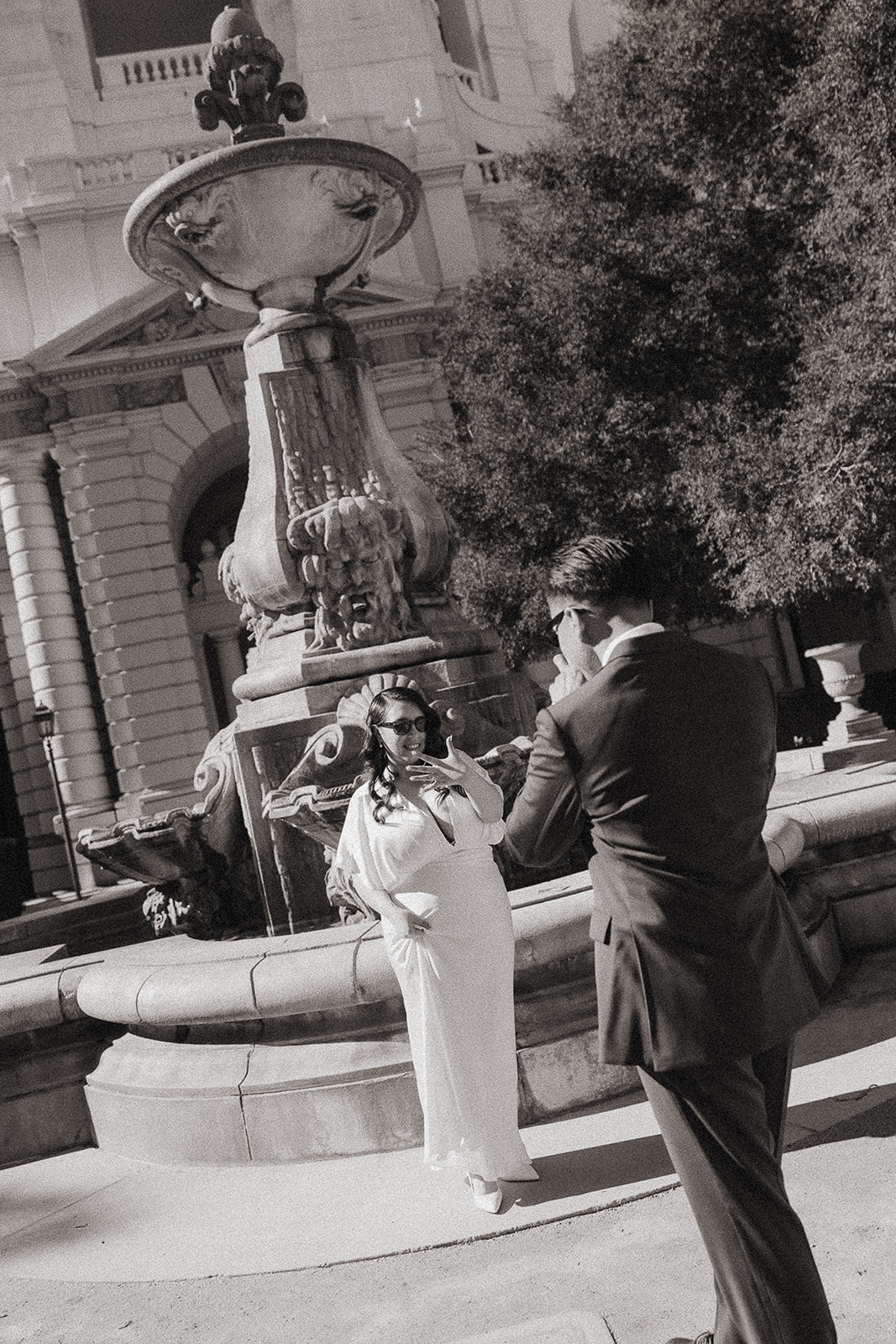 Black and white photo of the groom taking a photo of the bride in front of the fountain. 