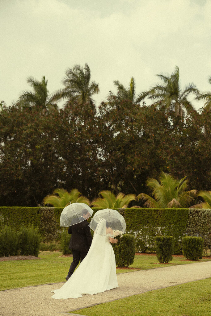 Far away shot of bride and groom walking away with umbrellas after their  Cinco Farm Gardens Miami wedding ceremony.