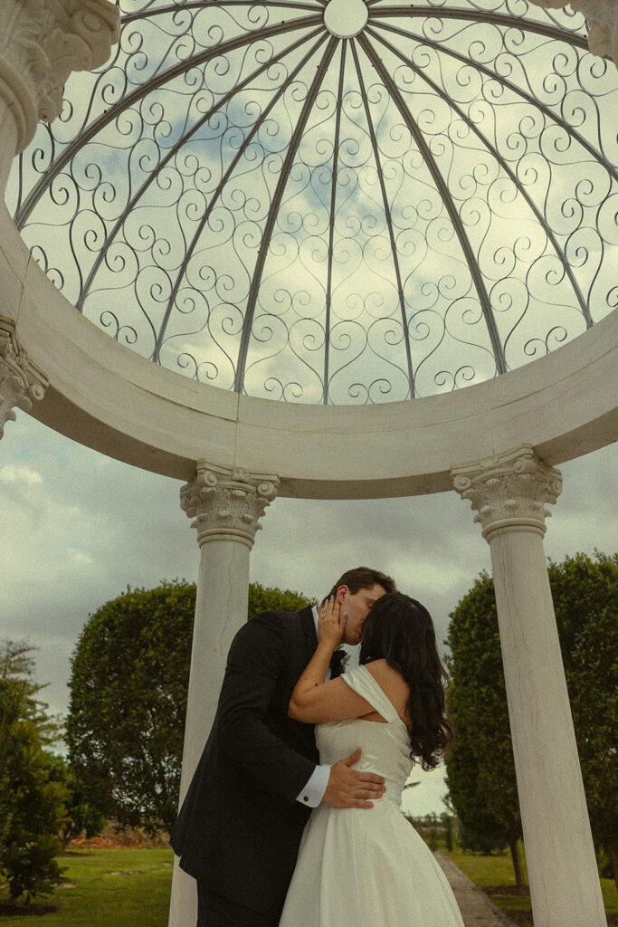 Bride and groom kissing under a white gazebo at Cinco Farm Gardens in Miami Florida. 