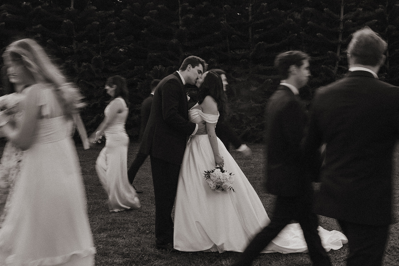Black and white motion blur photo of a bide and groom kissing as the bridal party walks by.