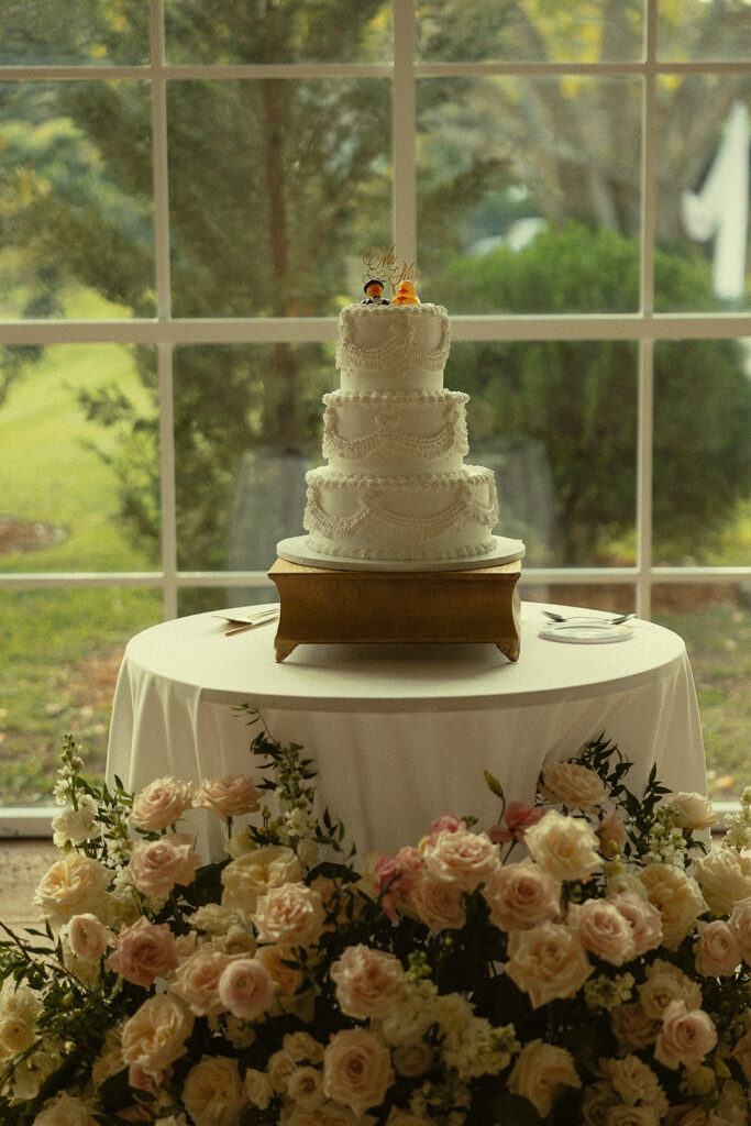 White three-tier wedding cake with floral decor by reception windows.