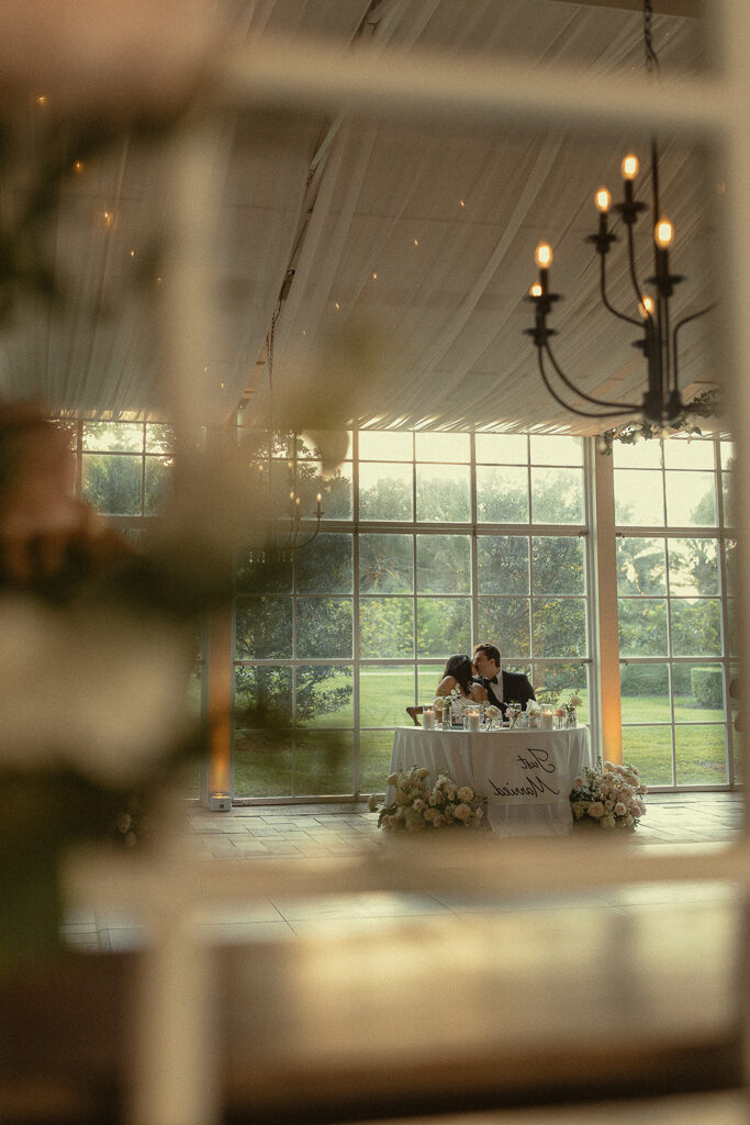Documentary style photo of a bride and groom kissing at their sweethearts table.