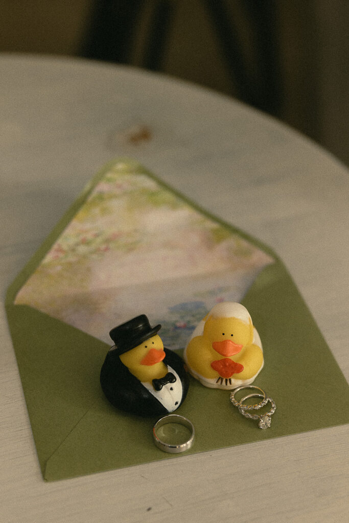 A bride and groom duck styled on top of a wedding invitation with rings. 