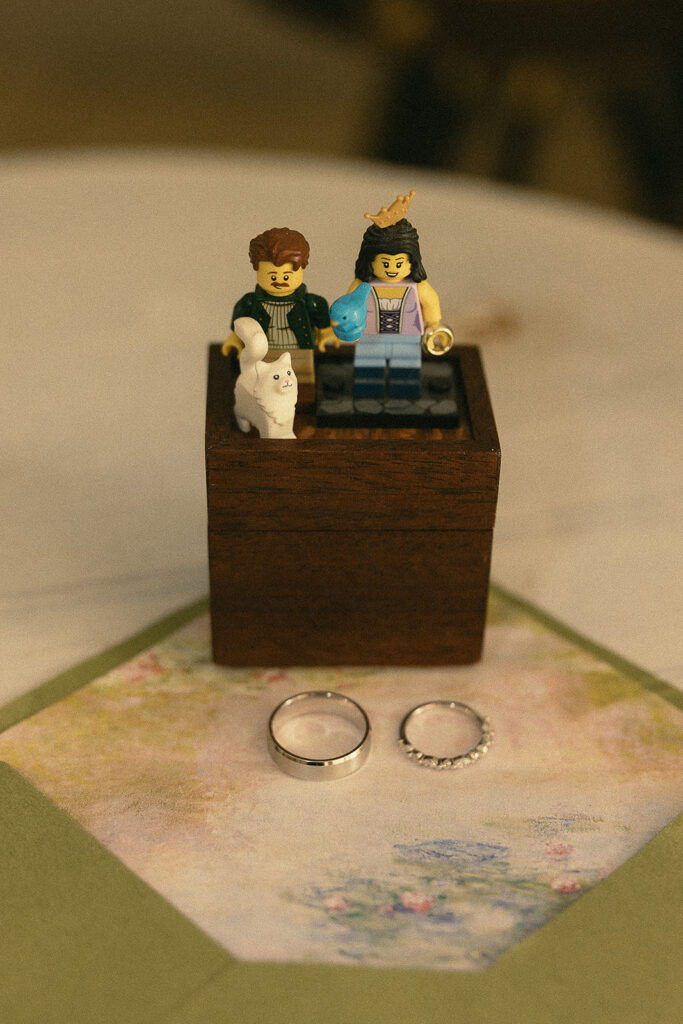 Lego figurines of a couple and a cat styled on top of their wedding invitation and rings.