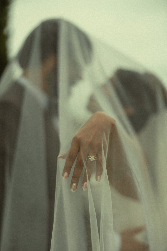 Bride and groom posing under the brides veil while she shows off her wedding ring.